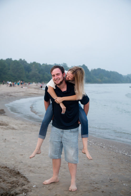 mangives fiance piggy back ride as she kisses his cheek port burwell engagement photography by london ontario photographer Woodgate Photography